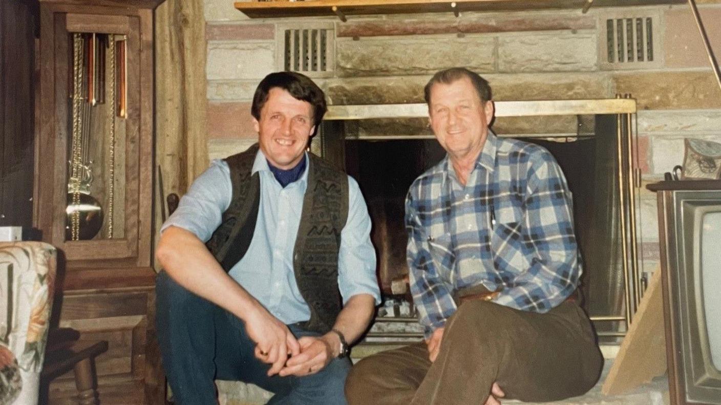 A younger Tim Lobb is wearing a gilet, a blue shirt and jeans. He is seated next to Marshall Donohew who is in a blue flannelette shirt and brown trousers. They are sitting in front of a fireplace.