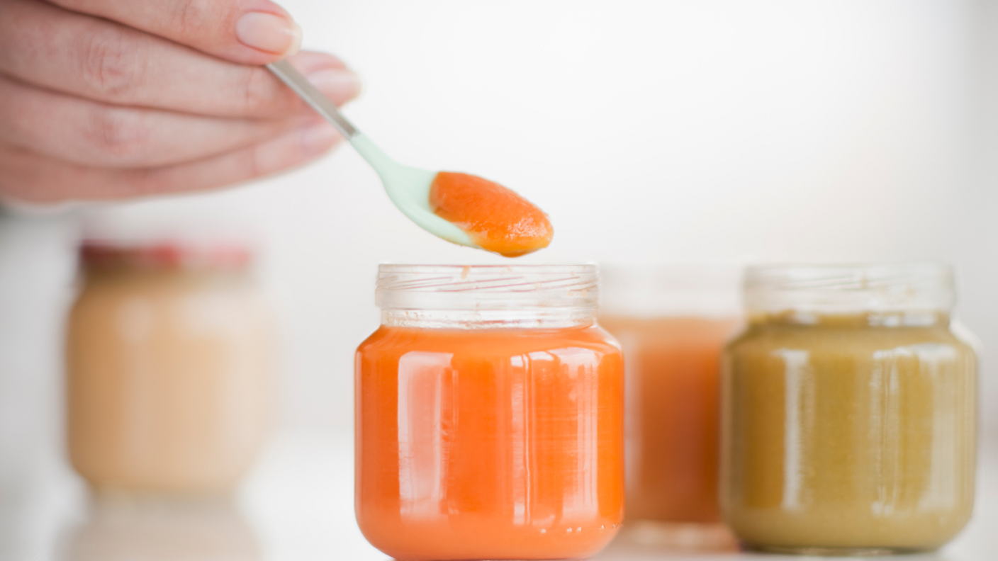 hand holding spoon with Orange coloured baby food, beside jars of Orange and brown baby food