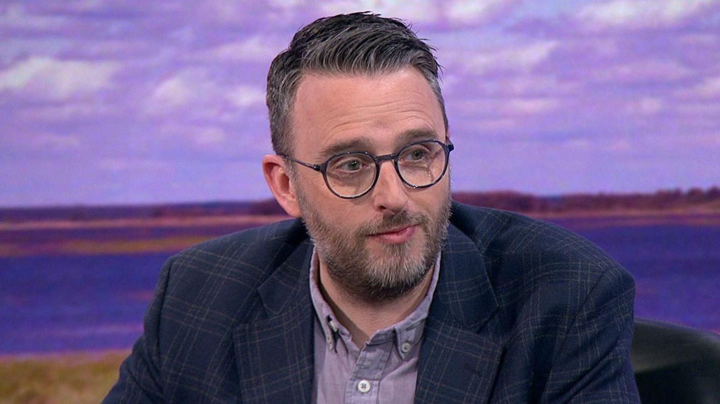 Nick Mathison, with dark hair and beard, wearing a checked blue jacket and purple shirt.