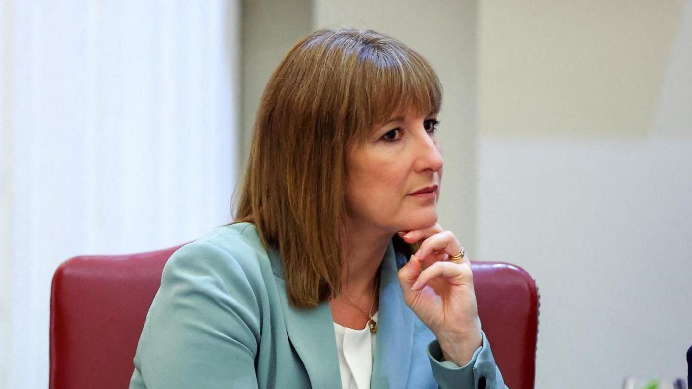 Rachel Reeves, wearing a turquoise suit and white top, sits in a red leather chair. Her hand is under her chin as she listens to someone speak.