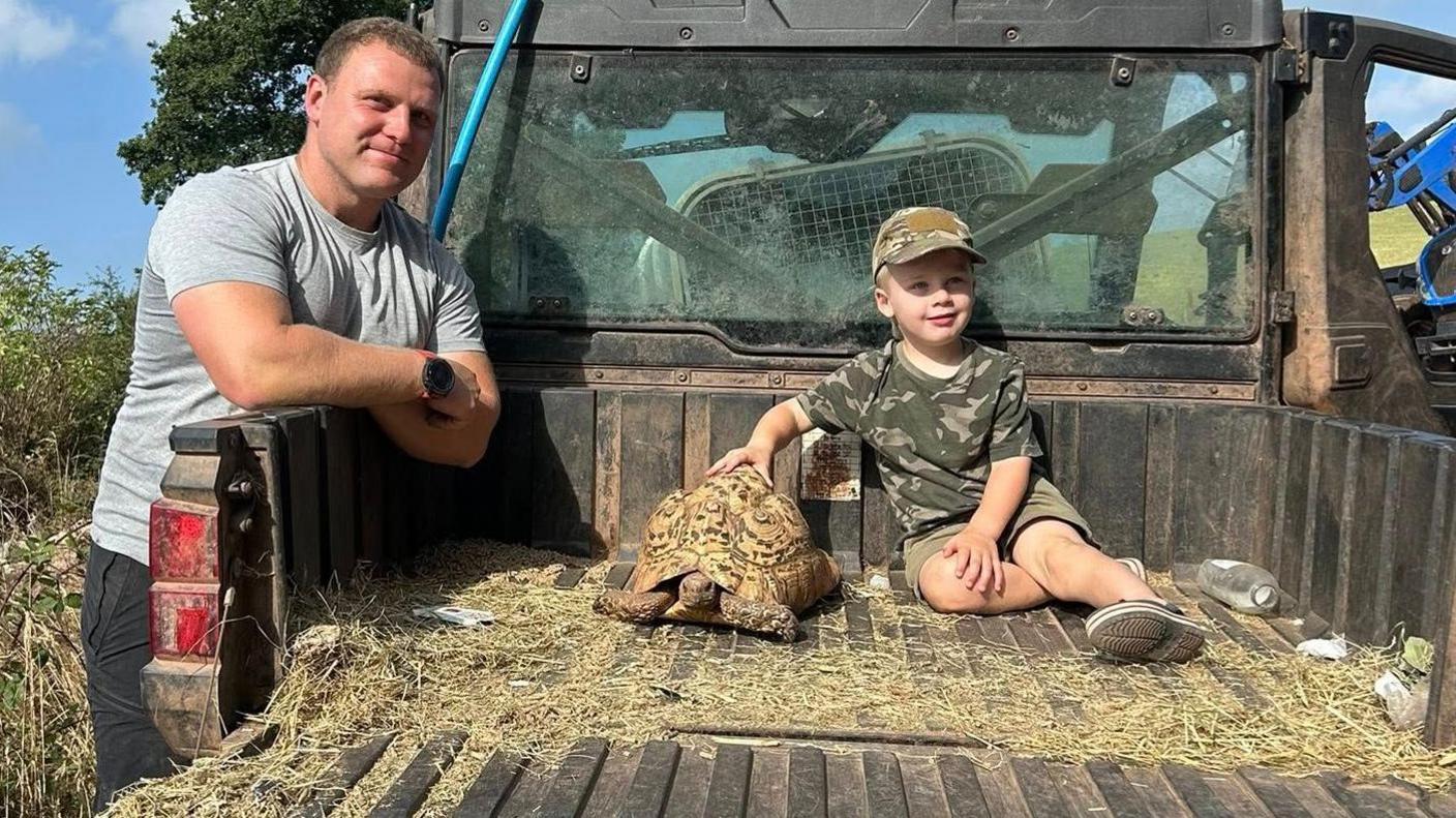 An adult in a grey T shirt with his arms over a pick-up where a young person sits holding a large tortoise with straw on the floor.