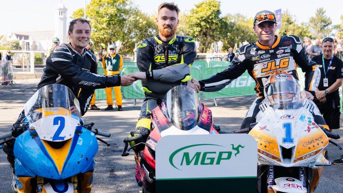 The top three riders sitting on their bikes shaking hands. All three are smiling.