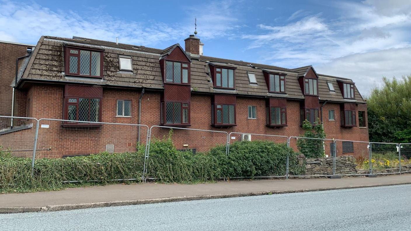 Metal fencing surrounding a stone wall overgrown with foliage in front of a red brick building behind with dark brown dormers.