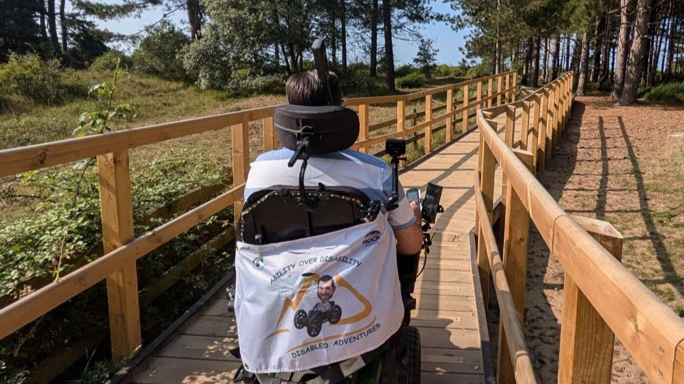 A man in a wheelchair moves away from the camera along a wooden boardwalk with wooden fencing on both sides preventing a small drop to sandy ground that is lined by trees.