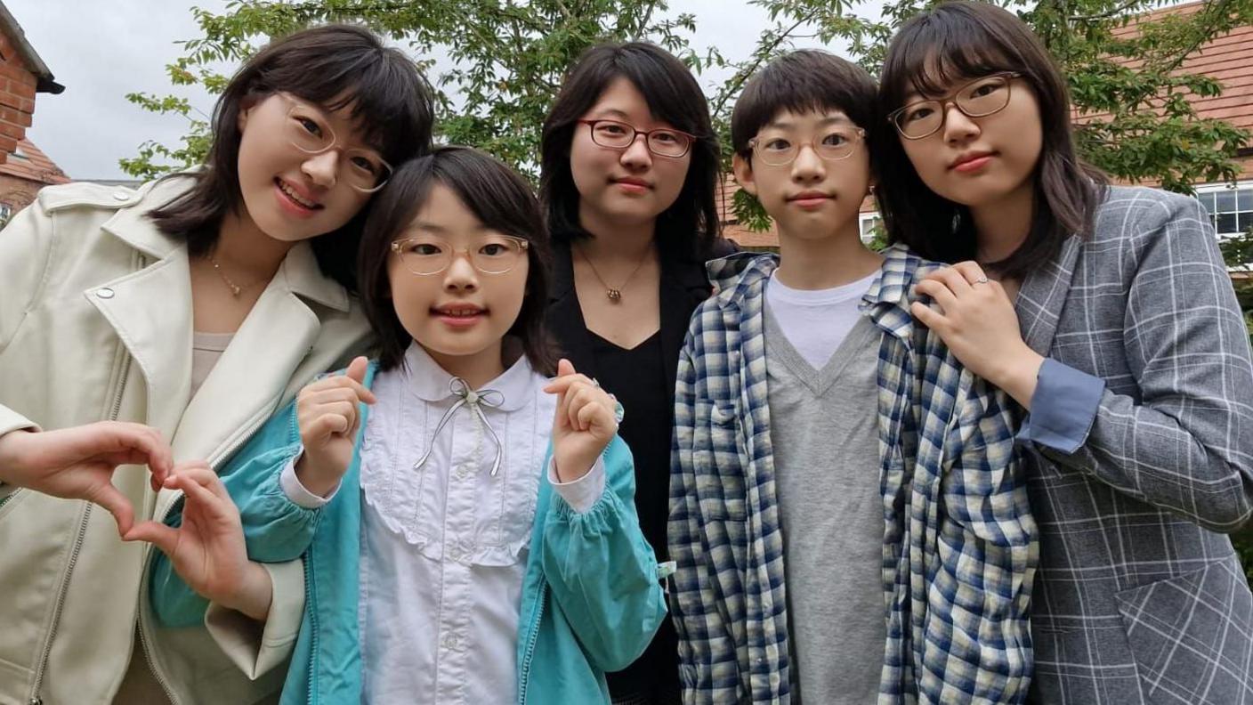 A young girl and a teenage boy stand in front of three young women. The girl has her hands up and is smiling. The woman on the left has her hands shaped like a heart. 
