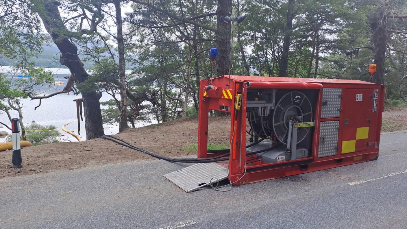 A large, red pump is sitting on a road, draining water out of a loch that is surrounded by trees  