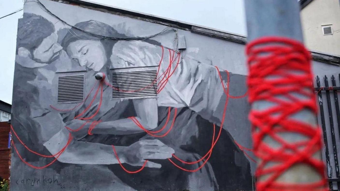 A black and white mural of a man and a woman in a cuddling position entangled in red threads. In the foreground red thread is wrapped around a street post and appears to be connected to the red threads on the mural.