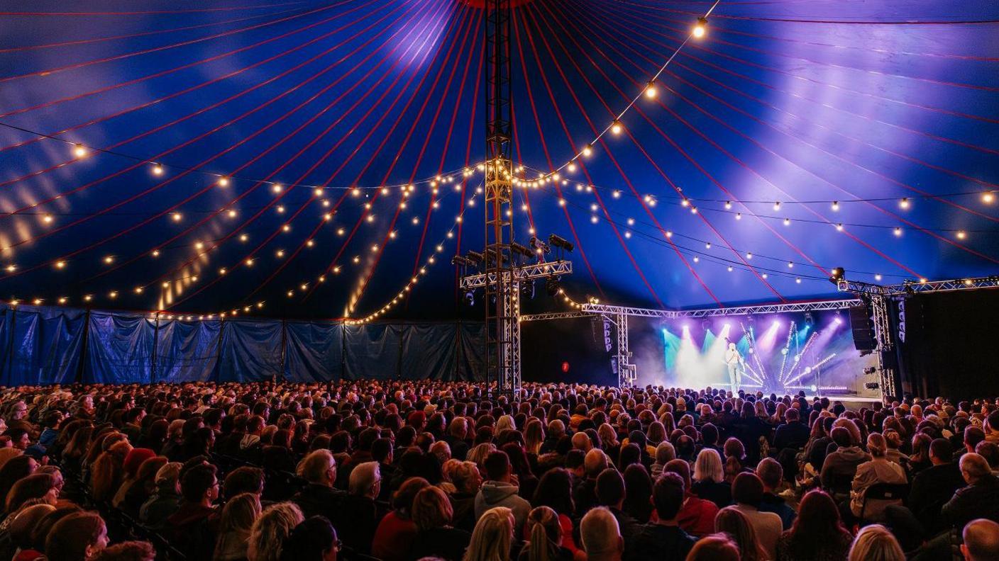 Thousands of people sit in the huge tent at Bristol Comedy Garden 2024. The image is a wide shot showing the entire tent plus the stage, which is illuminated by floodlights