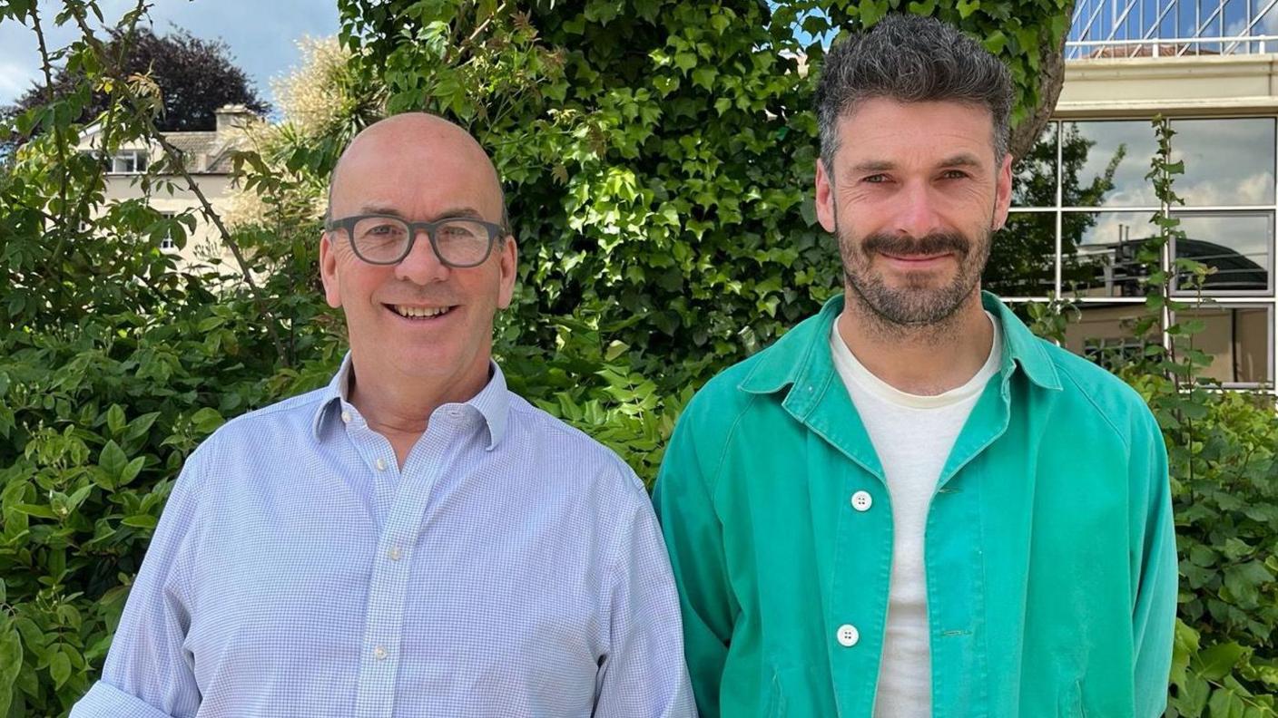 Sam Wells and Henry Herbert. They are pictured outside by a hedge. Sam is bald and has glasses with grey frames. He is wearing a white and blue check shirt. Henry is stood next to him. He has short dark hair and facial hair. He is wearing a turquoise shirt and white T-shirt. Both men are looking at the camera and smiling.