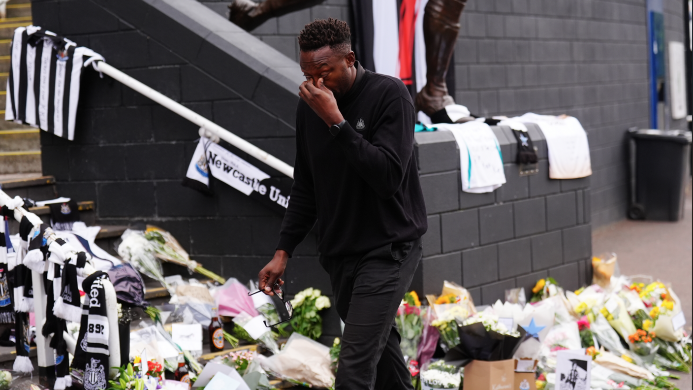Shola Ameobi, dressed in black, wipes away tears as he leaves St James' Park. The steps behind him are covered in flowers with Newcastle United shirts and scarves hanging from the banisters.