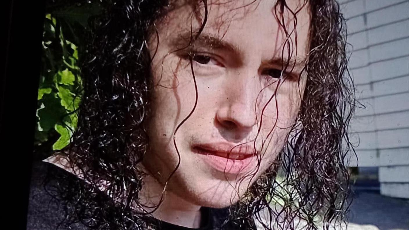 Vlad Nikolin-Caisley looks at the camera through his long, curly and black hair which hangs down to his shoulders. He is wearing a black t-shirt.