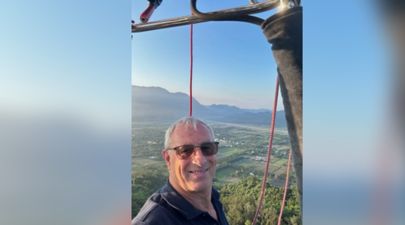Ian Martin pictured in a hot air balloon flying in the sky. He has short grey hair and is wearing dark sunglasses. He is looking at the camera and smiling.