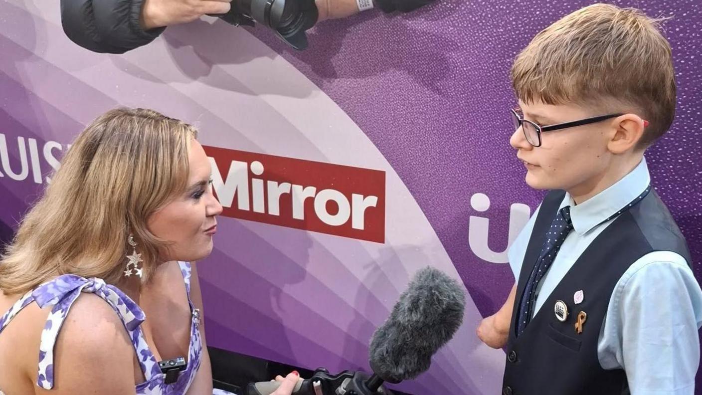Luke Mortimer is interviewed on the red carpet ahead of the Pride of Britain awards. Luke is wearing glasses and a black vest top and dark tie with a pale shirt. A woman is kneeling next to him holding a large microphone.
