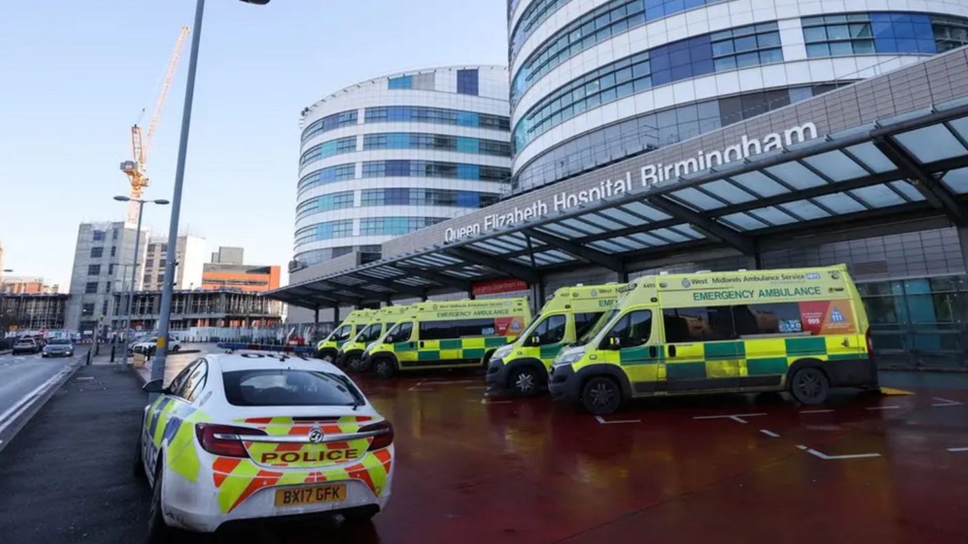 The front of a hospital building with a row of ambulances at the front and one police car. The sign above the door says Queen Elizabeth Hospital Birmingham