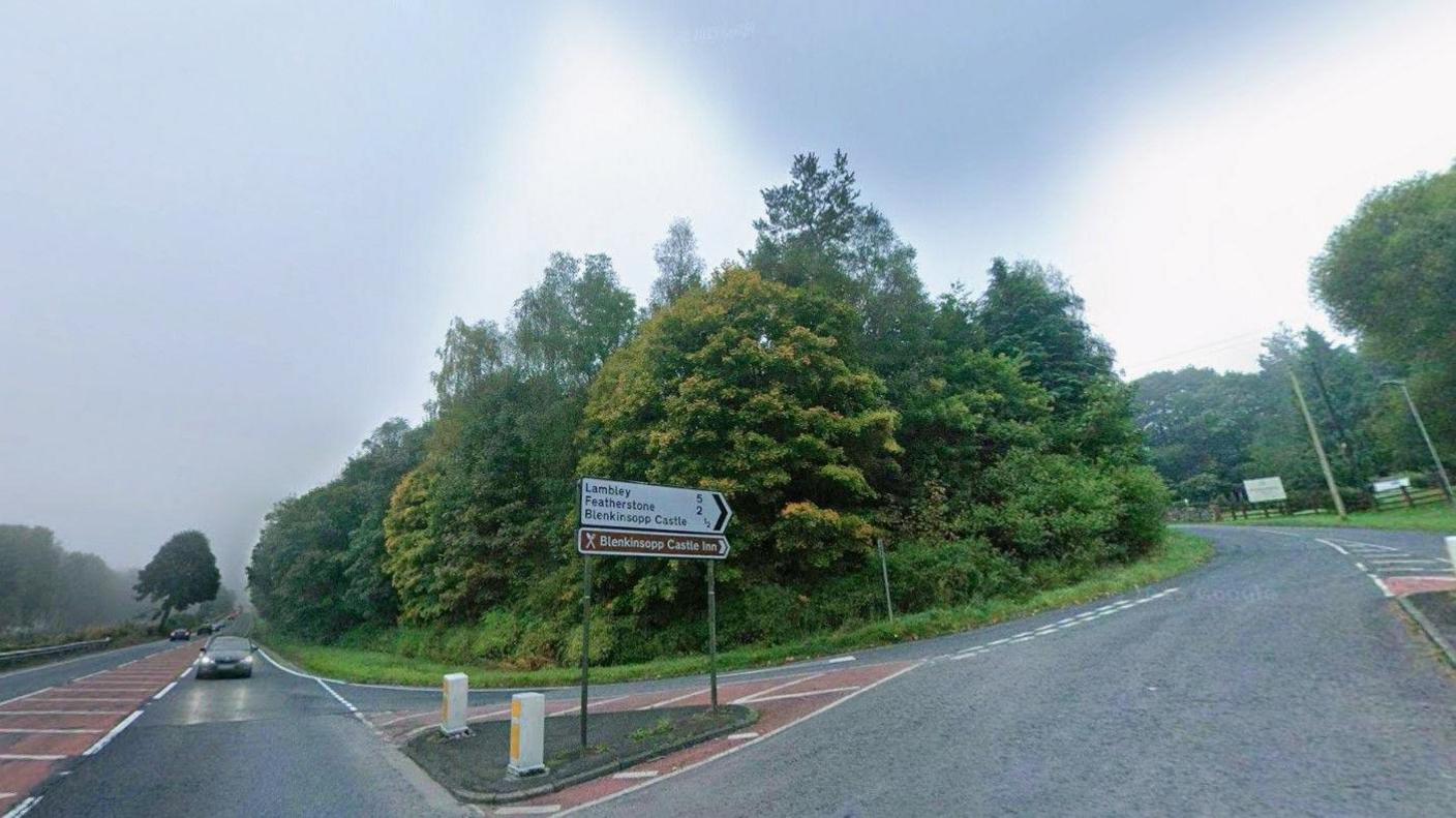 The Blenkinsopp Castle junction on the A69, close to where the fatal crash occurred. The A69 ploughs a straight course into the distance and has light traffic on it. Cars have their lights on as it is an overcast day and there is evidence of light rain. A road leads off to the right. It travels uphill then has a steep curve to the left which takes it behind a thick crop of mature trees. A white and black road sign points the way (with distances) to - from top to bottom - Lambley, Featherstone and Blenkinsopp Castle. Below that is a brown sign pointing to Blenkinsopp Castle Inn.