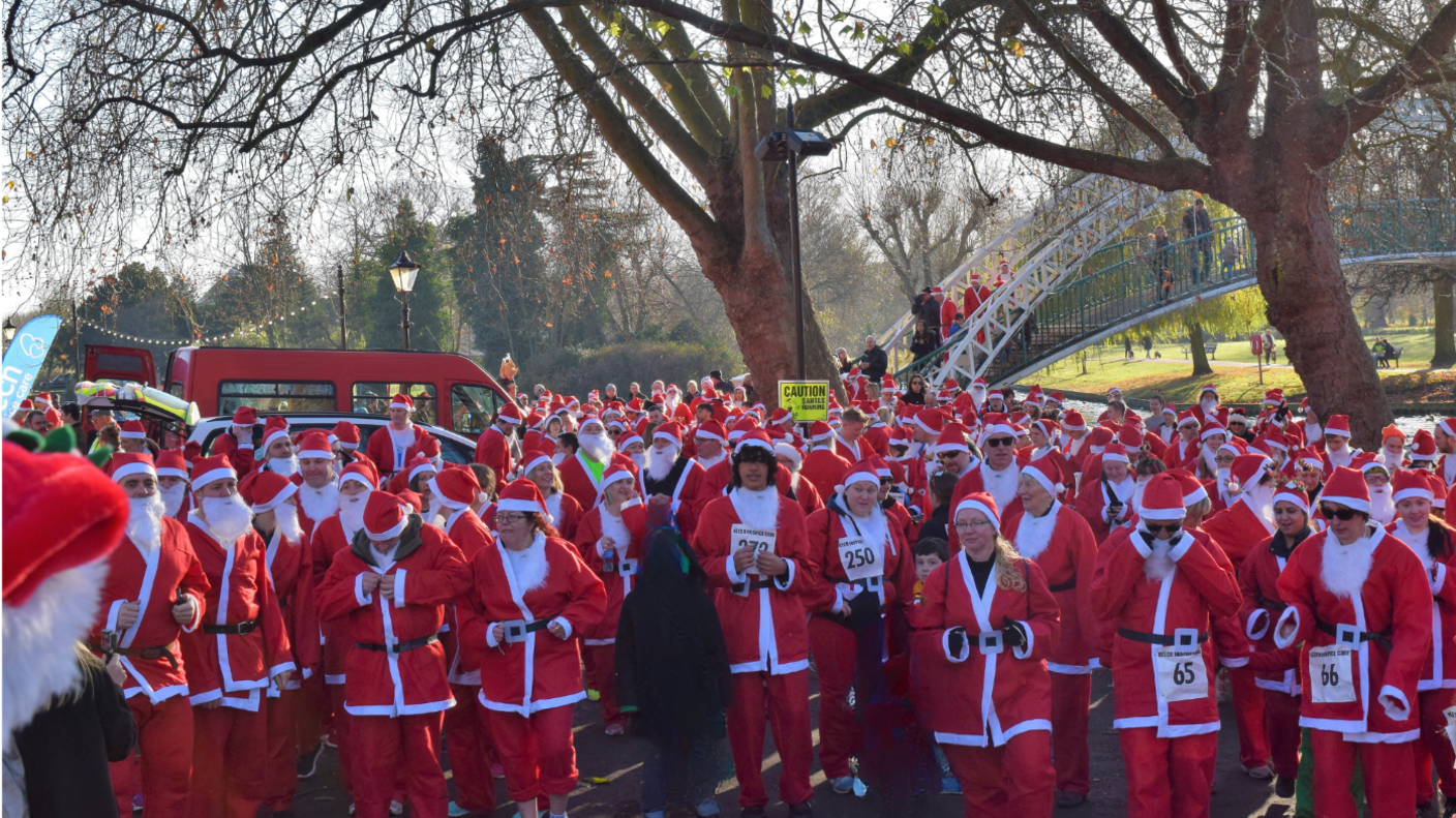 A large group of people wearing red Santa suits with hats. All the participants have numbers on them. There is a red van and a white bridge behind them