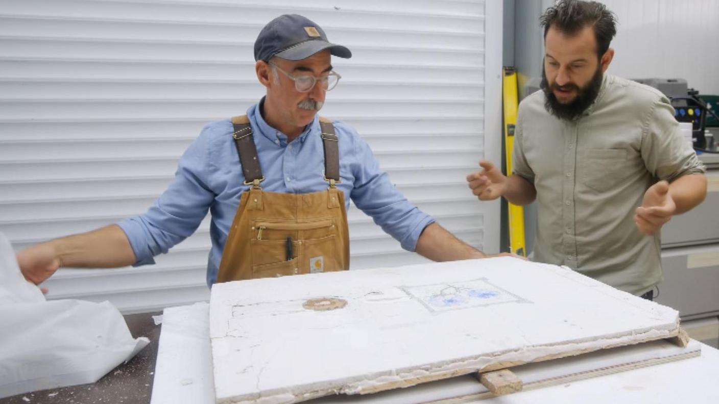 A man wearing a baseball cap, with grey hair and a moustache. He is wearing overalls and is looking at plaster. Next to him is a man in a khaki shirt with dark hair and a beard is is also looking at the plaster.