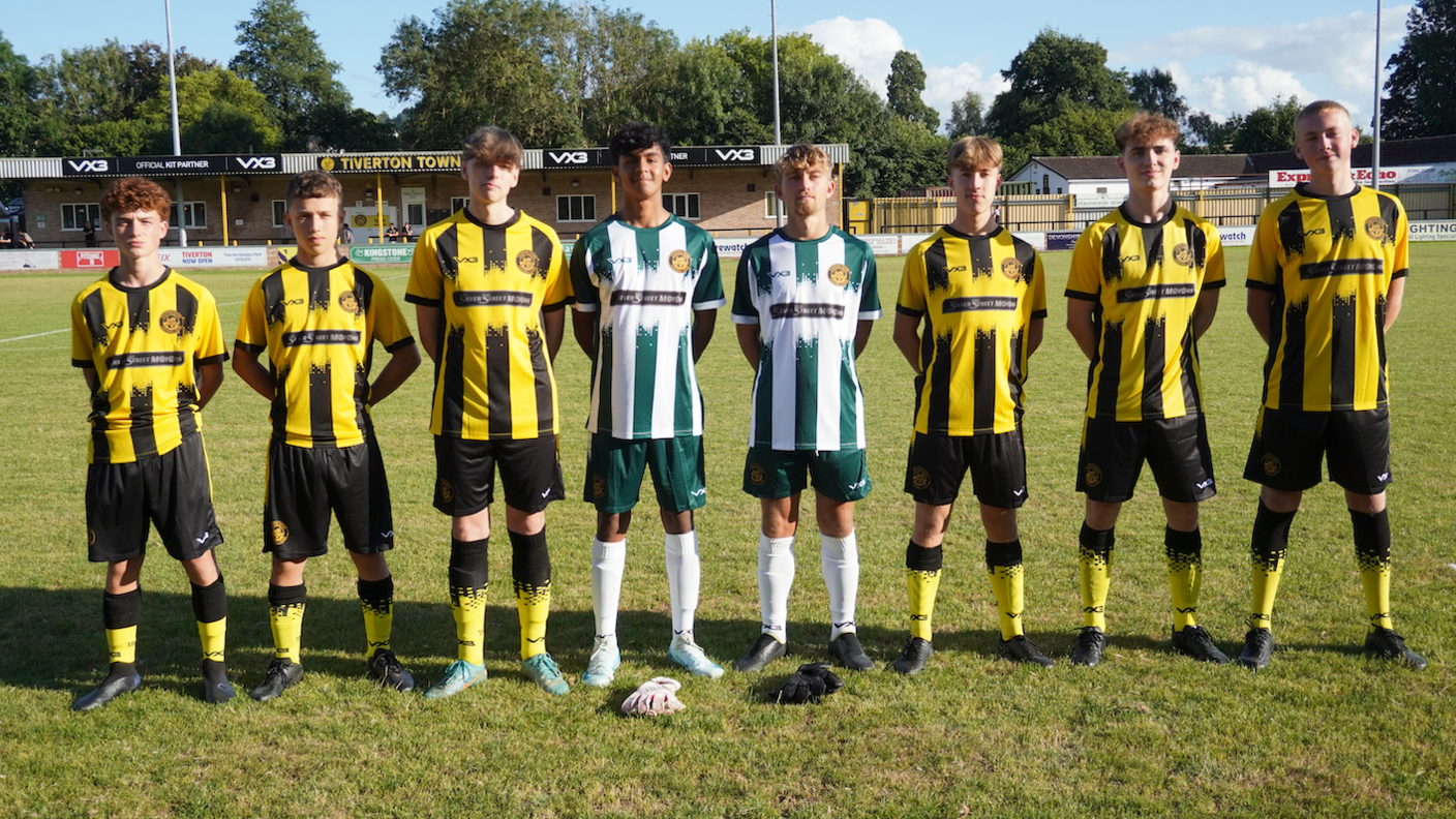 Eight under 18s standing in a line with hands behind their backs smiling on a football pitch on a sunny day