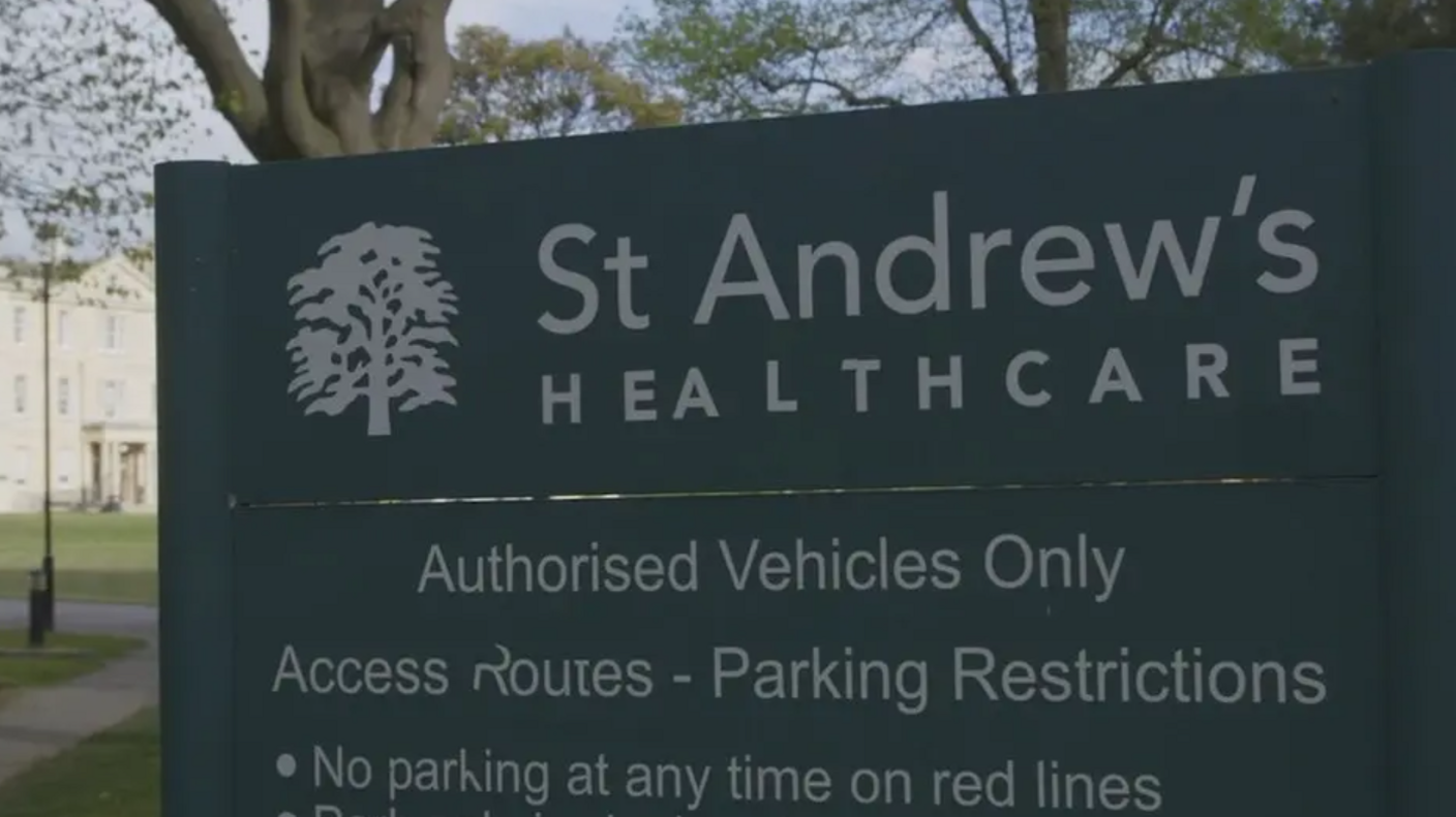 Sign at the front of a hospital. It is green with white letters. It says 'St Andrew's Healthcare' in prominent letters. behind the sign is a building.