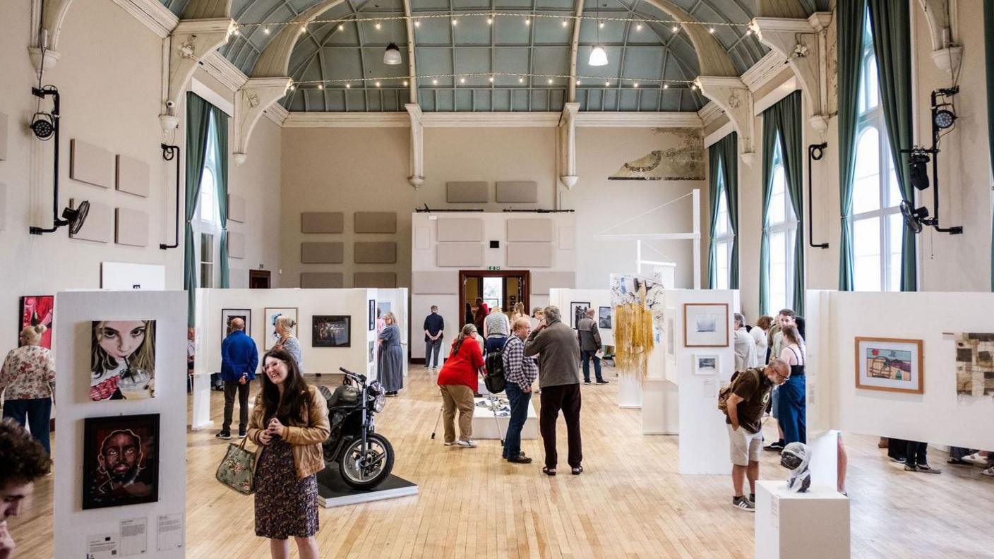 People view a selection of various pieces of artwork, including paintings and photographs, which on display as part of an exhibition. A motorbike can also been seen on display. 