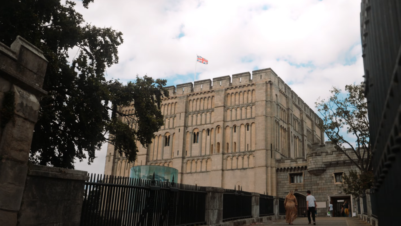 Norwich Castle, and the bridge leading up to it. The sky above it is blue but cloudy. In the foreground, there is the walkway to the door of the castle. There are two people walking across the walkway.