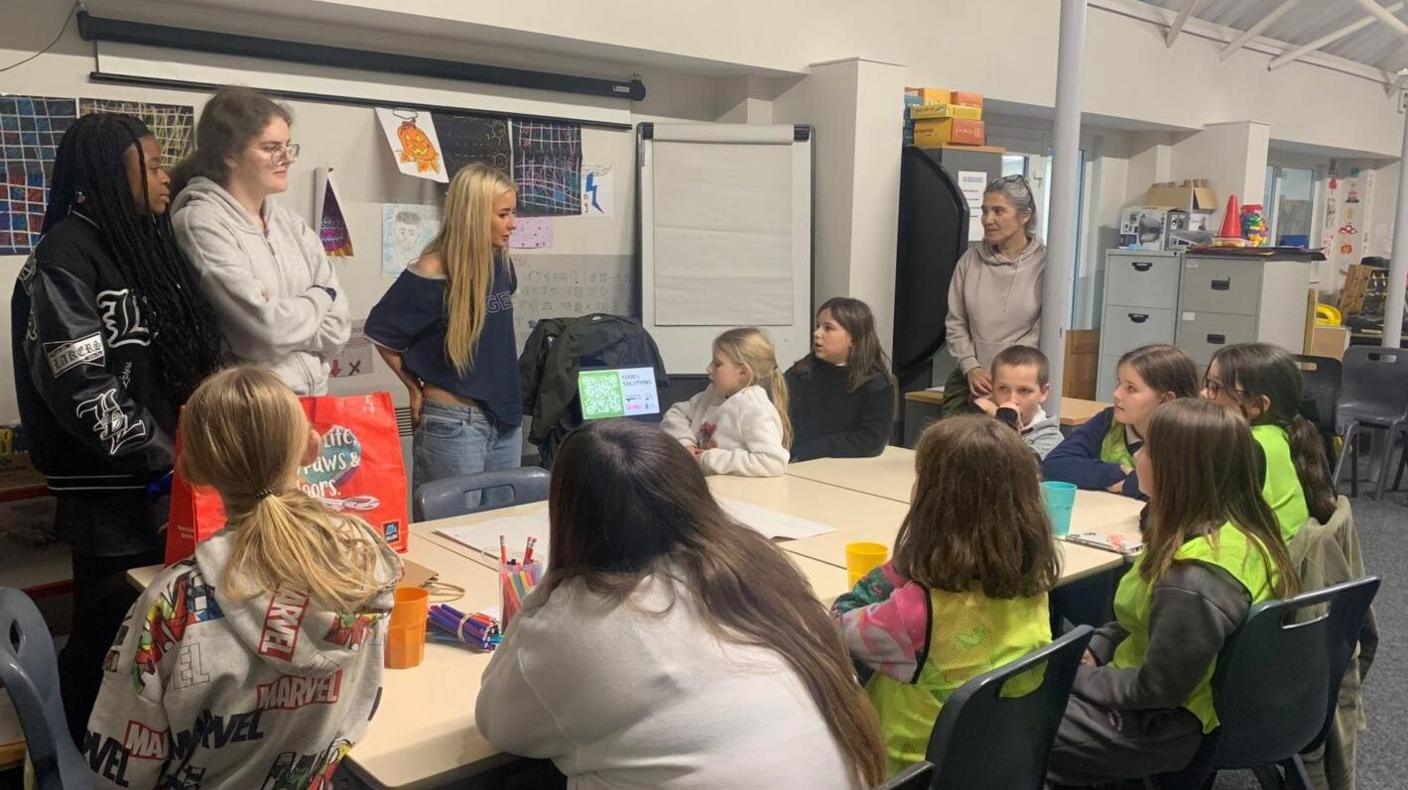 A woman and 13 children sitting around a table looking at a presentation on a laptop.