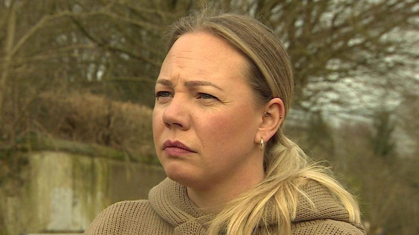 A woman with blonde hair, which is in a ponytail, wears a beige hoodie, and has an expression of deep thought as she looks to the left of the camera, standing in a field next to a coffee van