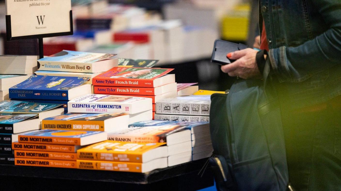 A small table in a Waterstones bookshop stacked with books and a person holding a phone and a bag, standing at the table