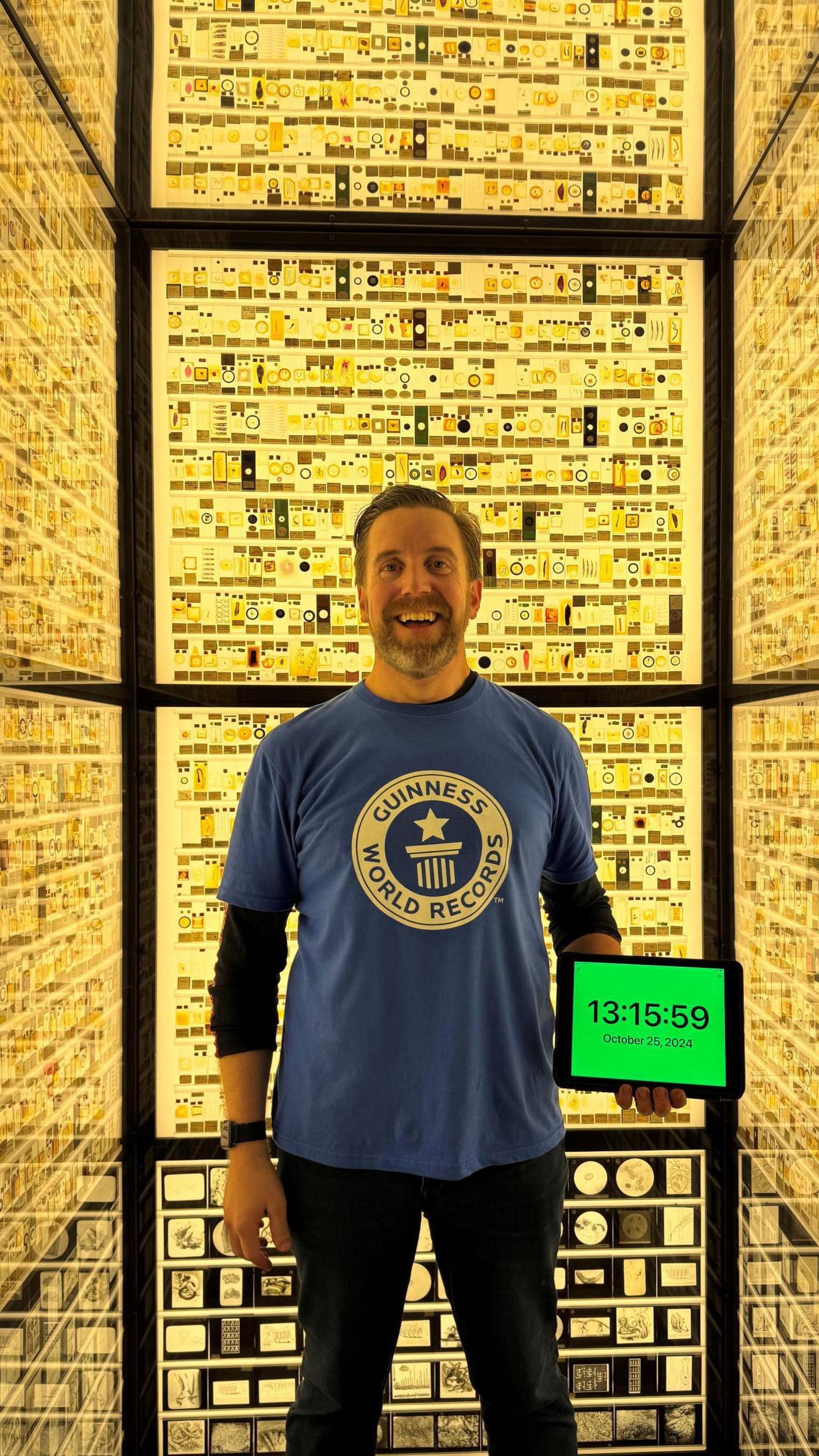 Ben Melham in a museum with lots of pictures. He is looking pleased and is holding a tablet with the timing on, as proof he has made the record.