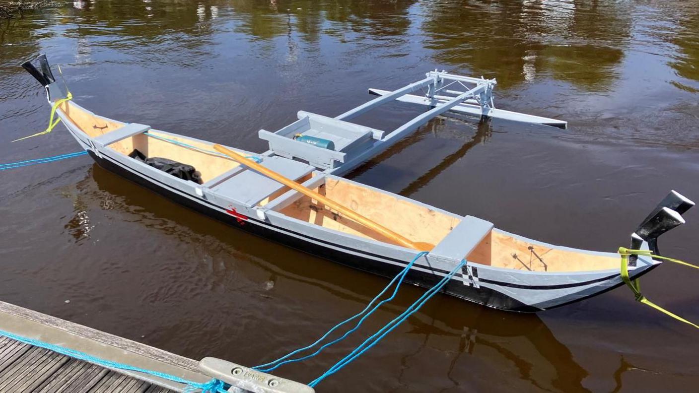 A wooden canoe, built by Micronesian boat builders sits on the water's edge. It is tied to a jetty with blue rope