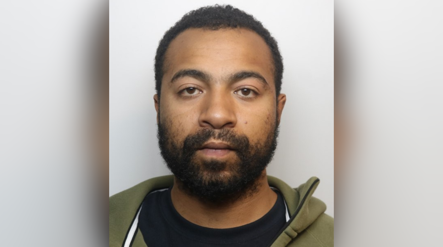 A custody shot of a man. He has short dark hair and facial hair. He is looking directly at the camera with a blank expression on his face. He is wearing a black T-shirt and green hoodie.