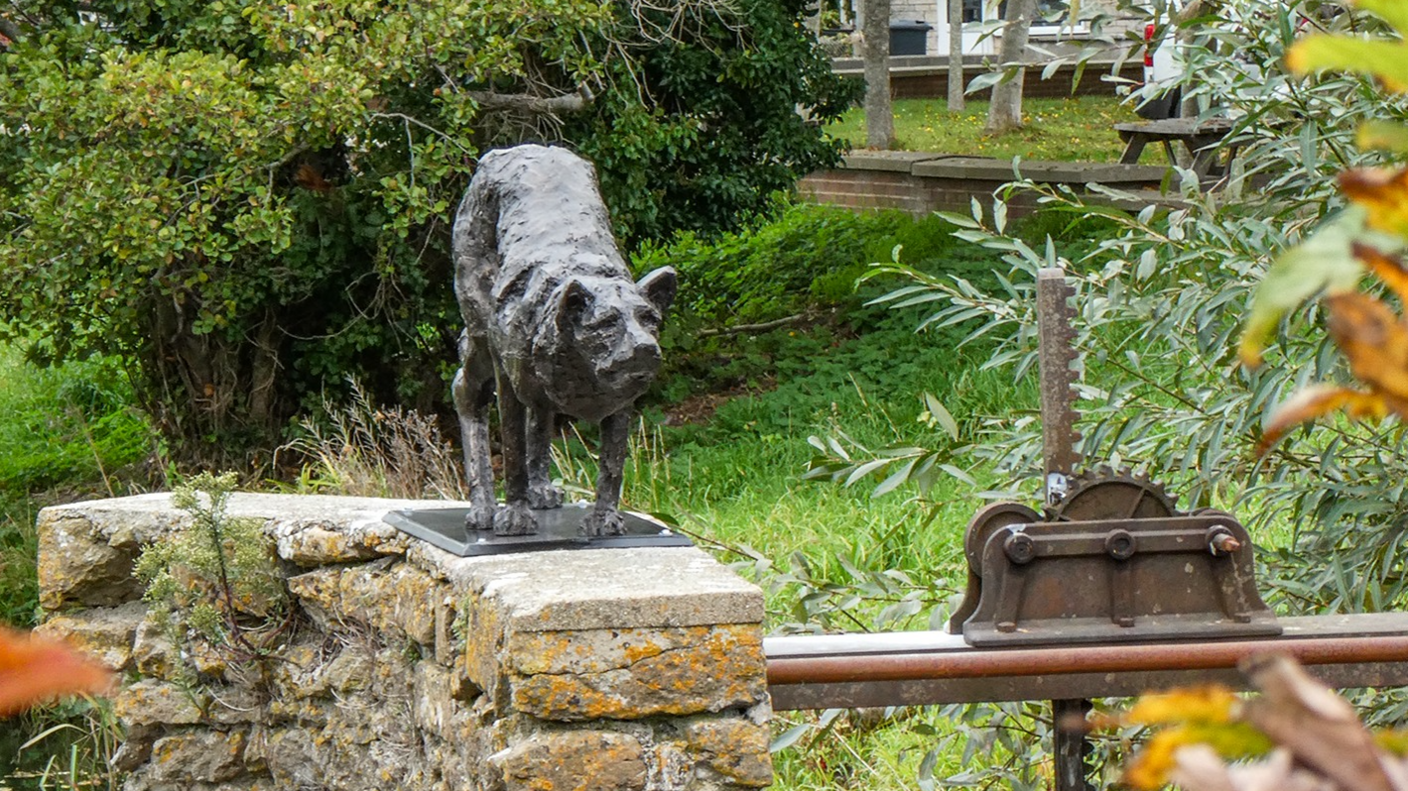 Front view of a black sculpture of a stalking dog on a stone wall next to a weir mechanism. The dog resembles a border collie when it is stalking sheep - its head is low and its back is raised with its tail tucked in.