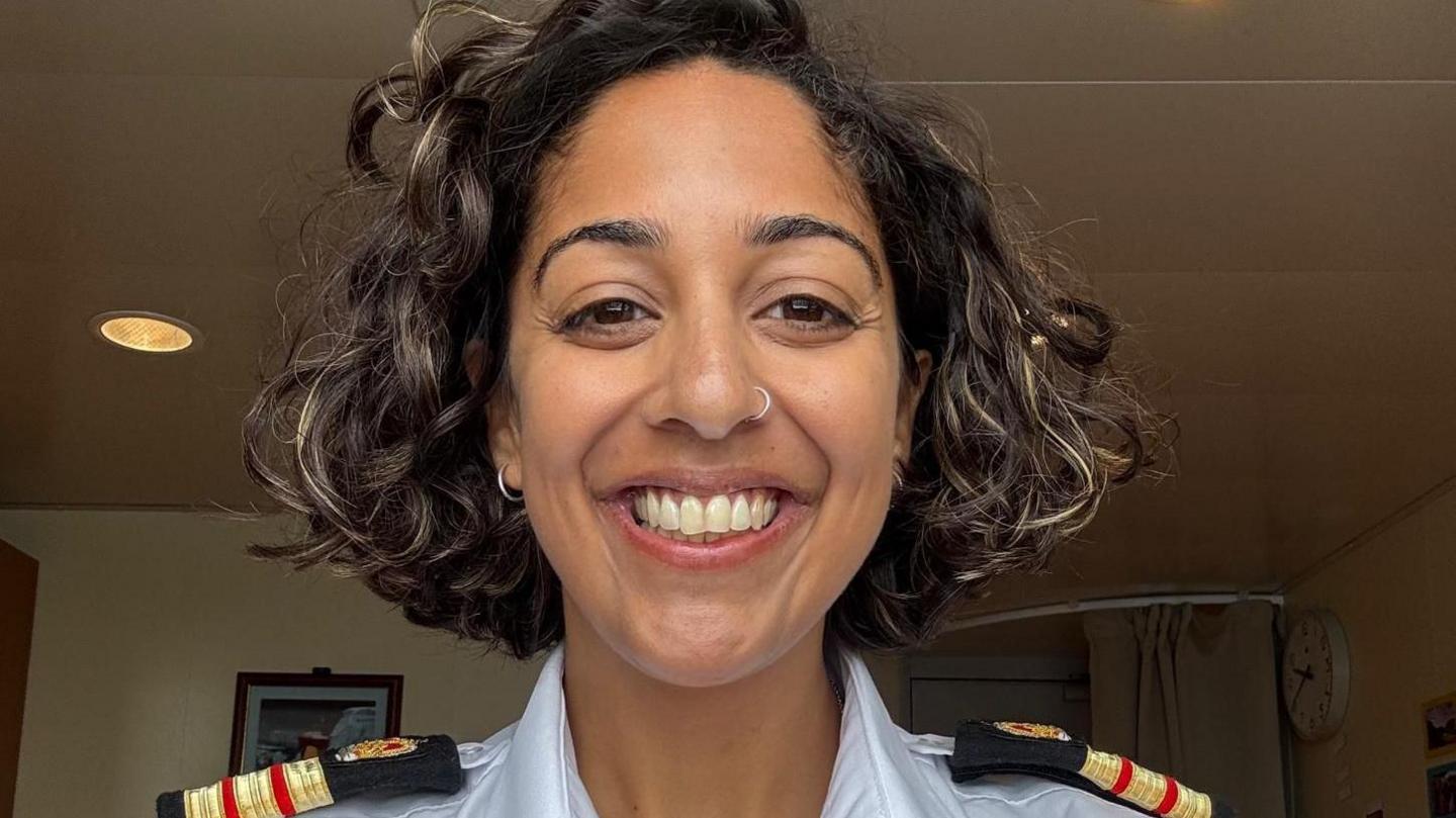 Dr Nisha Mistry is smiling at the camera. She has dark, wavy hair just above shoulder length. She is wearing earrings and has a ring through her nose. She is wearing a white shirt with epaulettes