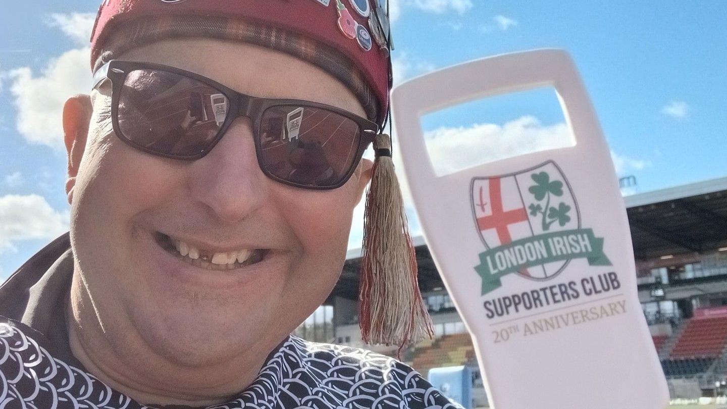 Nige Eaton in a fez and sunglasses, posing next to a London Irish Supporters Club 20th Anniversary cut-out sign