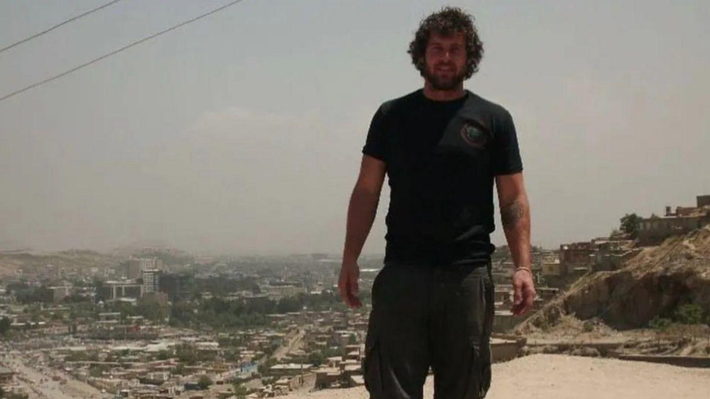 Paul Harris wearing a black t-shirt and trousers standing on a hill in Afghanistan with a town and mountains in the background