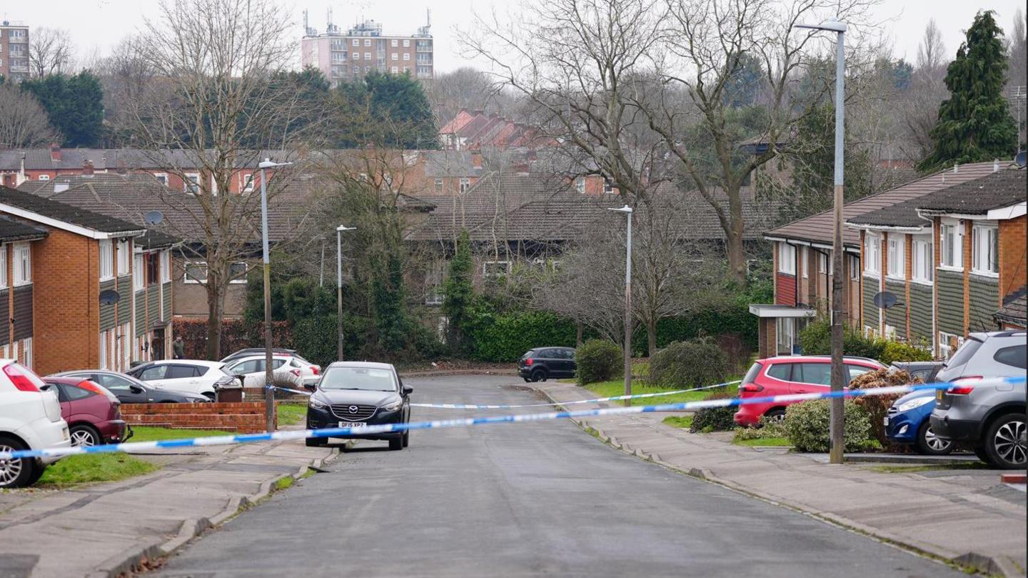 A residential street with cars and houses either side and trees in the background. A police cordon is in place across the middle of the street.