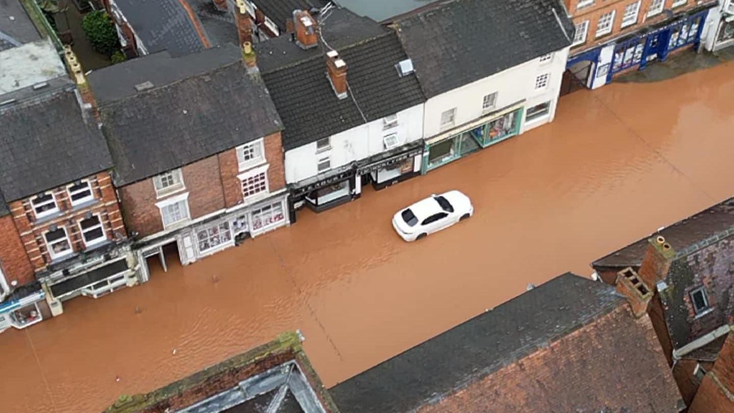 Flooded roads after the river bursts its banks in Tenbury Wells during Storm Bert. The water is brown and has flooded several streets and has flowed into houses and shops across the town. A white car can be seen on the road with waters nearly over its tyres.