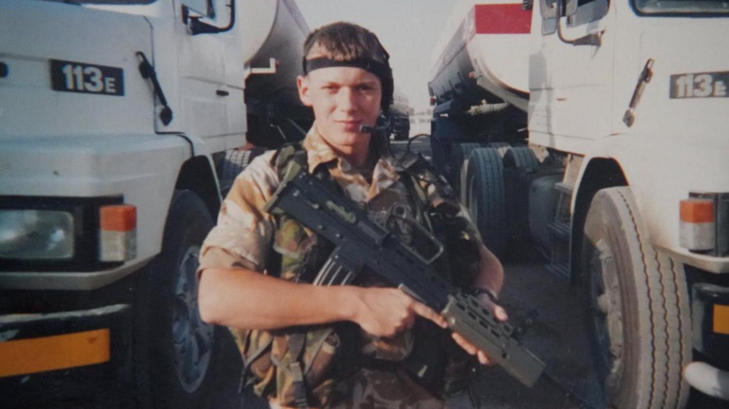 Rikki Drury as a young man in camouflage uniform holding a rifle across his chest. He is stood between two large lorries.