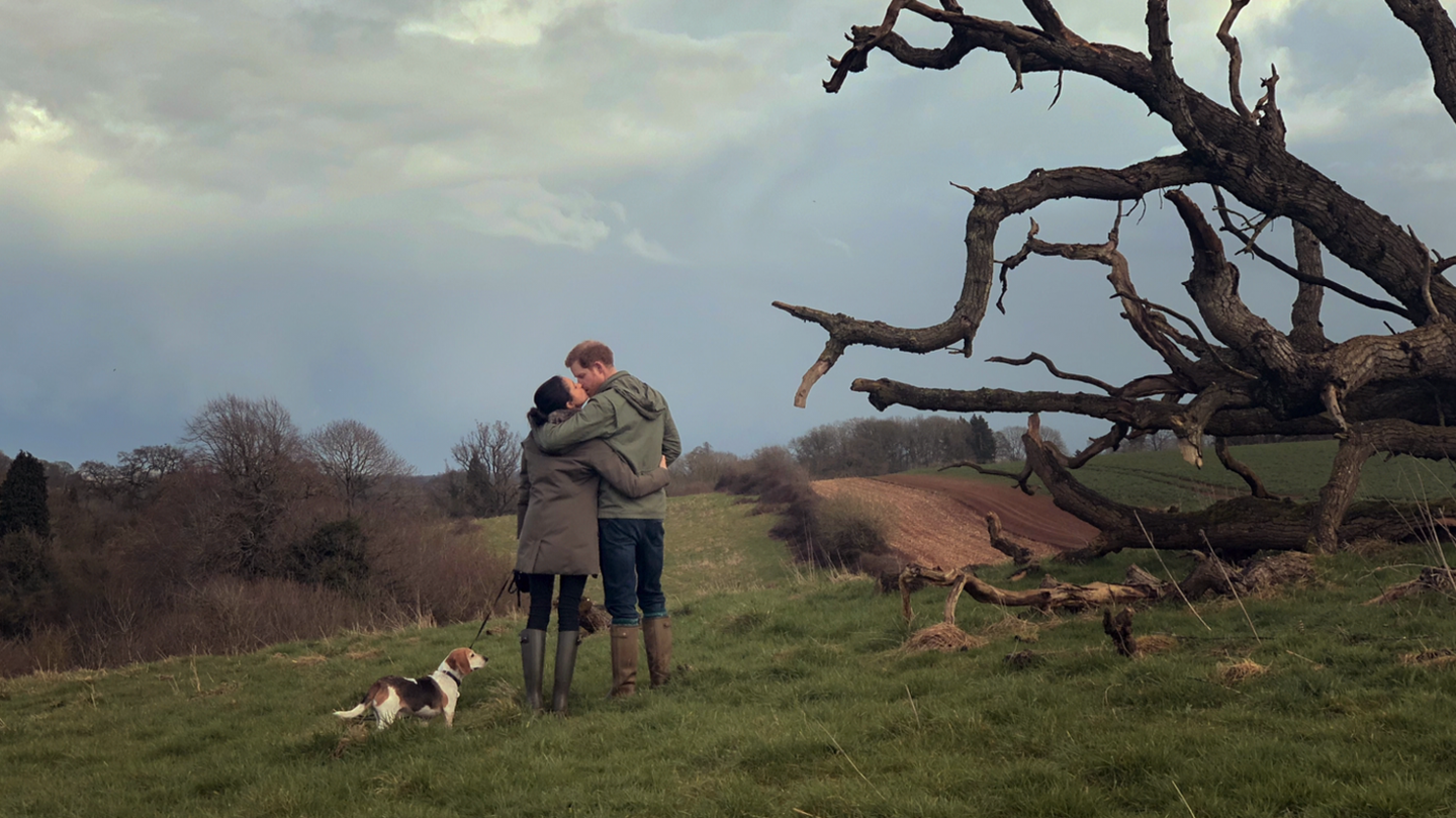 A handout photo shows the Duke and Duchess of Sussex with their late dog, Guy. The two are standing in a field in the middle distance with their backs to the camera. The duke's left arm is round the duchess's shoulders and the two are kissing. Guy is on a lead held by the duchess and is standing next to them. 