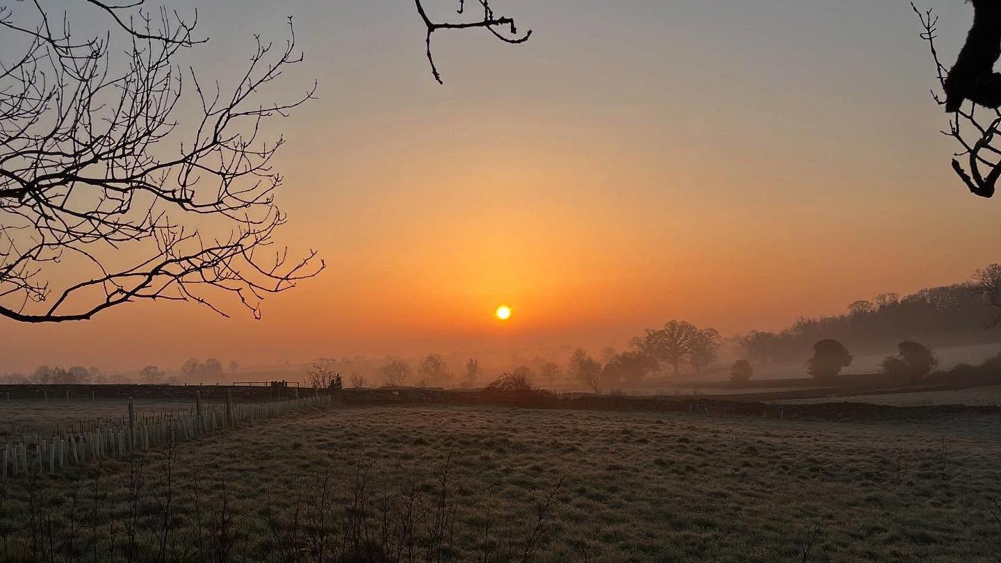 A sunrise with frosted fields in the foreground in the Cotswolds. Lines of hedgerows and trees are dimly visible in the haze and the sun is bright yellow with an orange haze around it