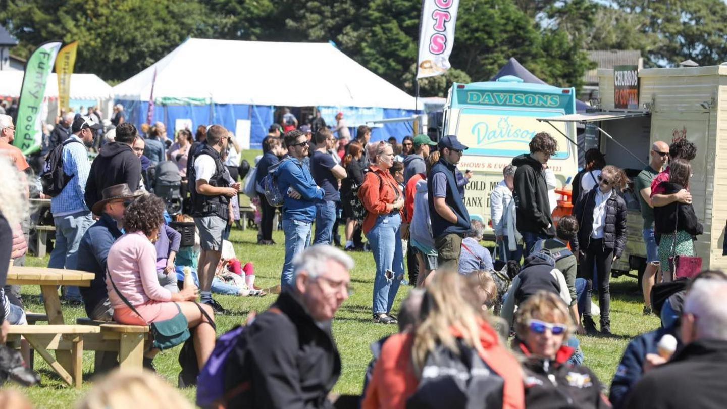 Crowds queuing for food, and people sitting down on benches in a grassed area on a sunny day. The stalls include a light blue and cream coloured Davison's ice-cream van.