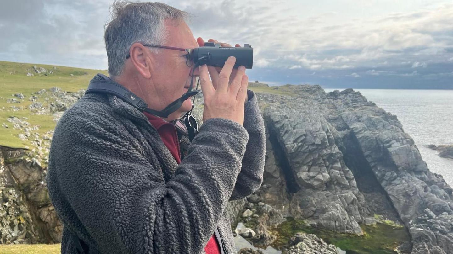 A man with greying hair and wearing a grey fleece jacket and red jumper. He is wearing glasses and holding binoculars to his eyes. He is standing on cliffs. More cliffs and the sea can be seen in the background. 