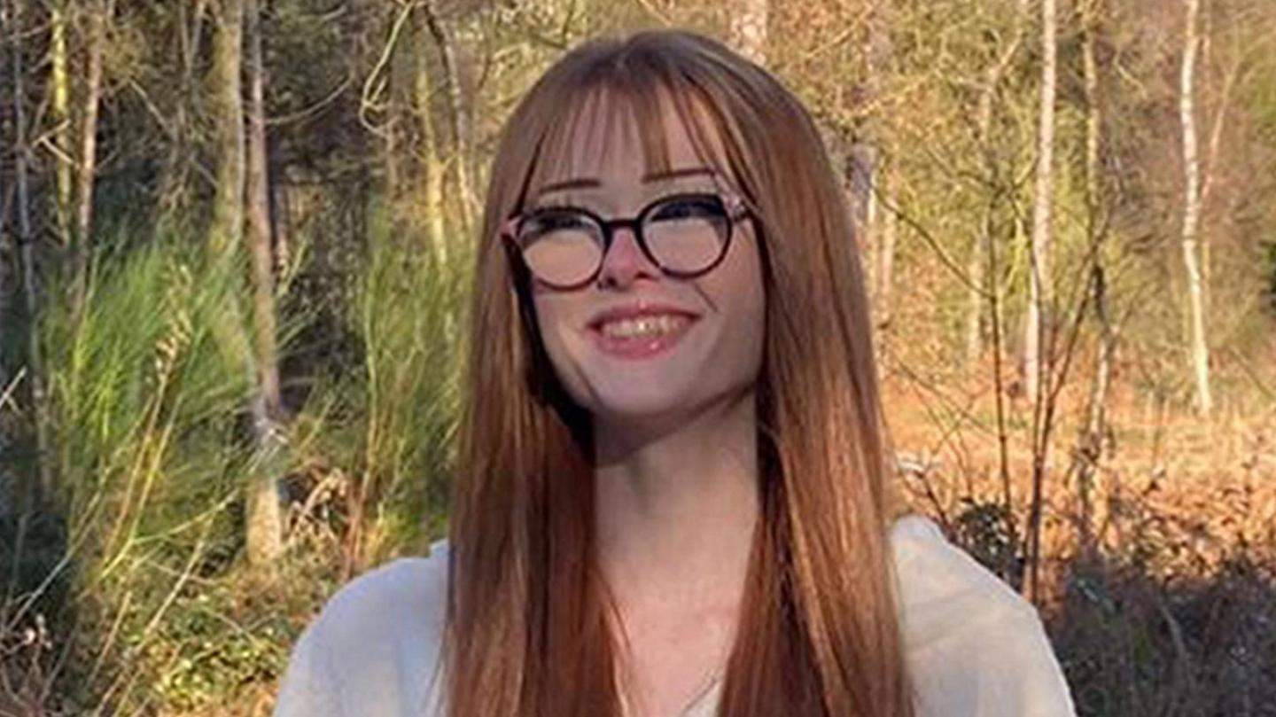 Brianna Ghey, with long red hair and glasses, standing with forestry behind her 