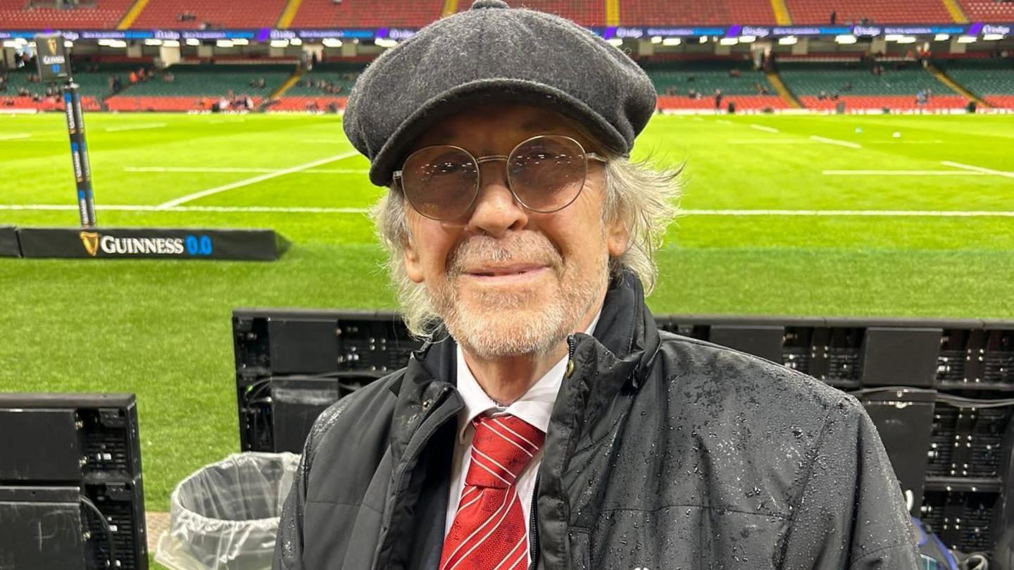 Iestyn Garlick yn gwenu at y camera gyda chae'r stadiwm yn y cefndir a seddi gwag. Mae e'n gwisgo sbectol, tei coch, cap fflat a chot ddu.