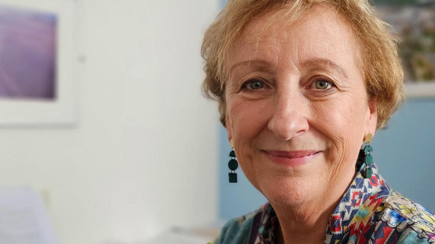 Group ceo Lesley Dwyer looks at the camera. She is wearing a multi coloured blouse, with dangly green earrings. The picture shows her head and shoulders, she is in her office. She has short blonde hair.