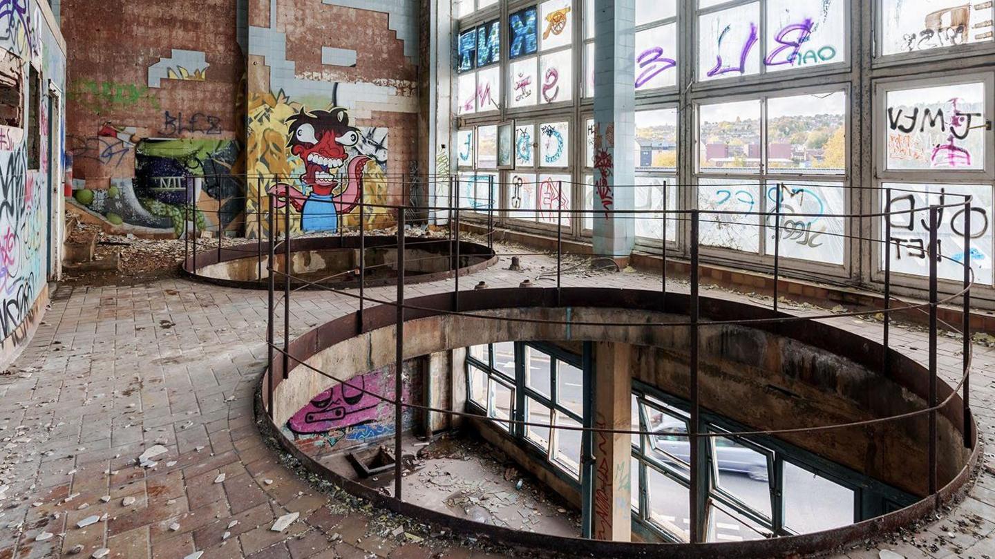 Inside a derelict building. A full-wall glass window is covered in graffiti on the inside and outside. There are tiles falling off the walls and dust from the degrading building. Mostly graffiti tags but on the far wall is a painting of a red octopus type monster.