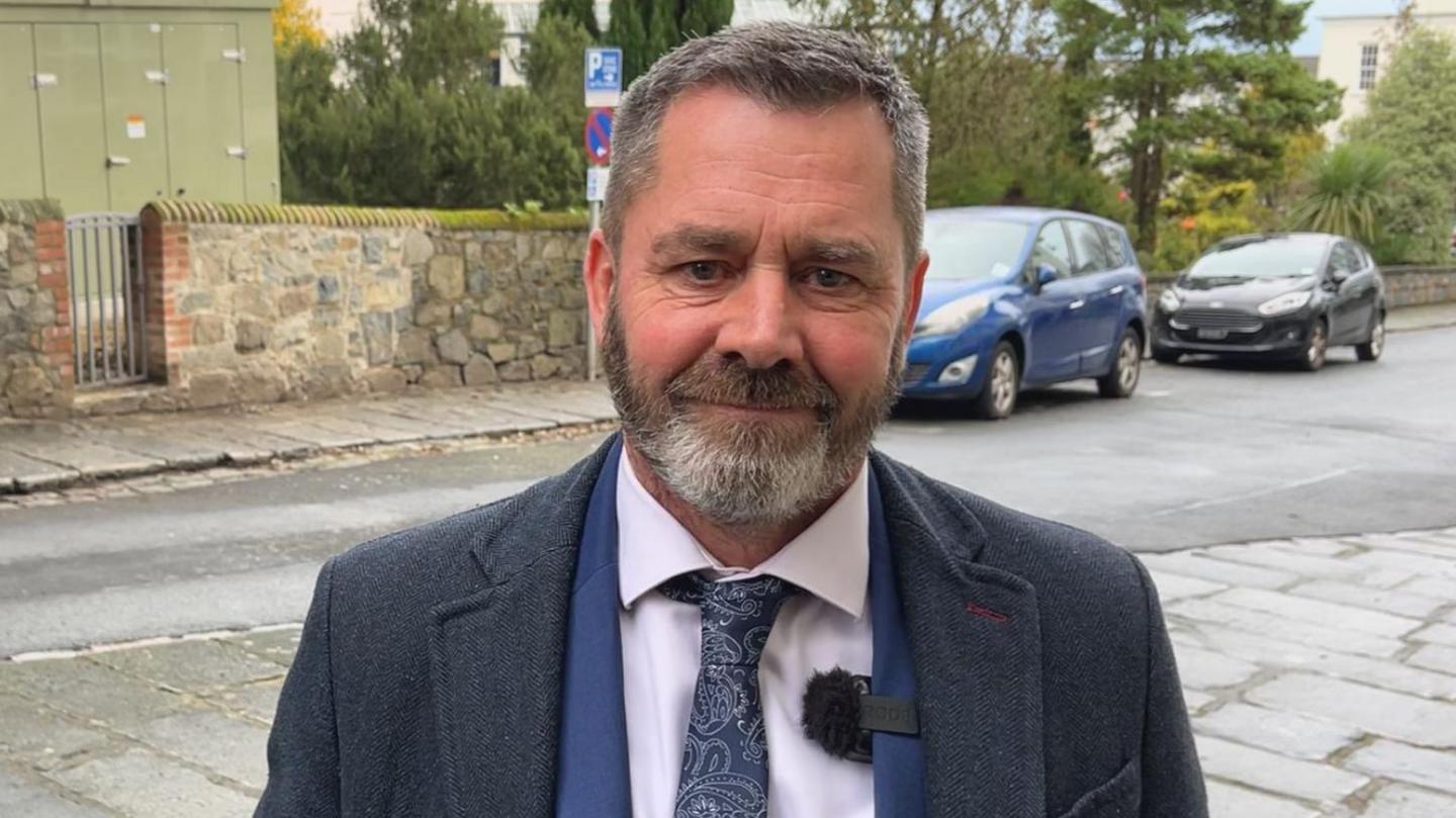 Deputy Marc Leadbeater - A man with grey and black hair and a grey and black beard. He's wearing a blue coat, a blue suit jacket and a blue patterned tie. On his lapel is a mic fluffy. 
