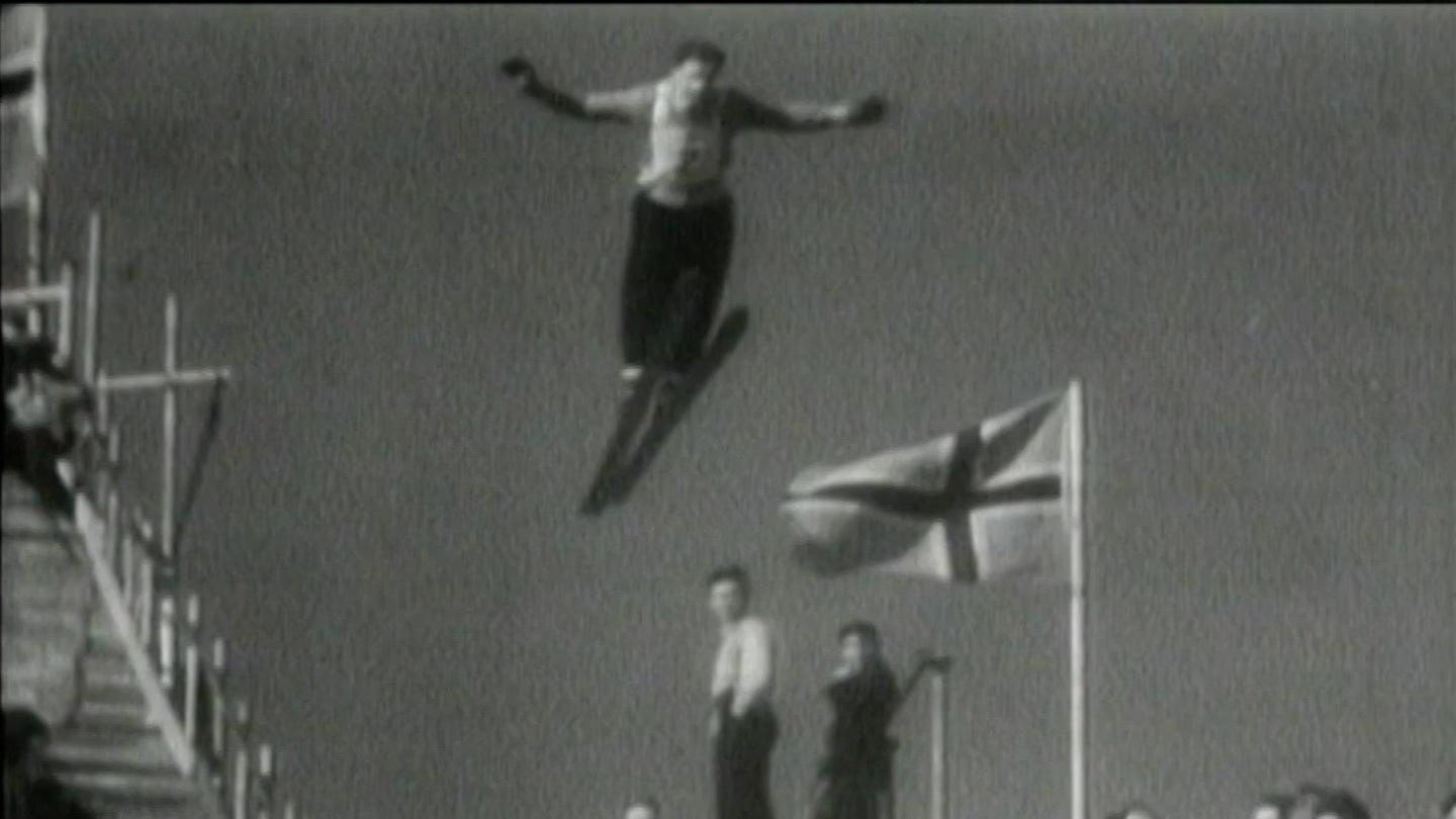 A black and white photo of a ski jumper taking off and flying through the air, with his arms outstretched. A Norwegian flag is fluttering on a flagpole behind him.
