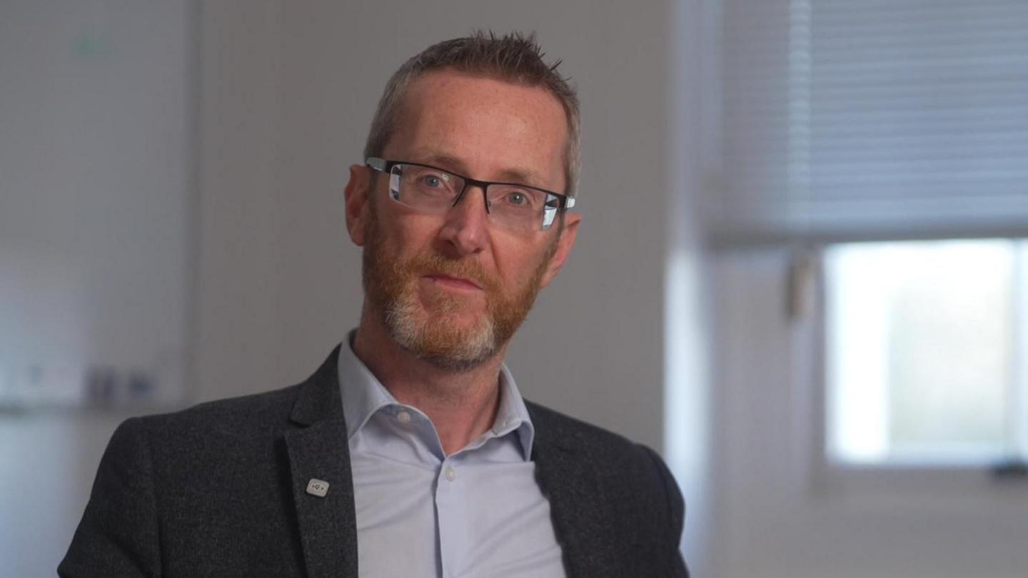 A man with short, dark hair spiked at the front. Wearing black glasses, a grey blazer and blue shirt. He is pictured against a cream wall with a window behind him.