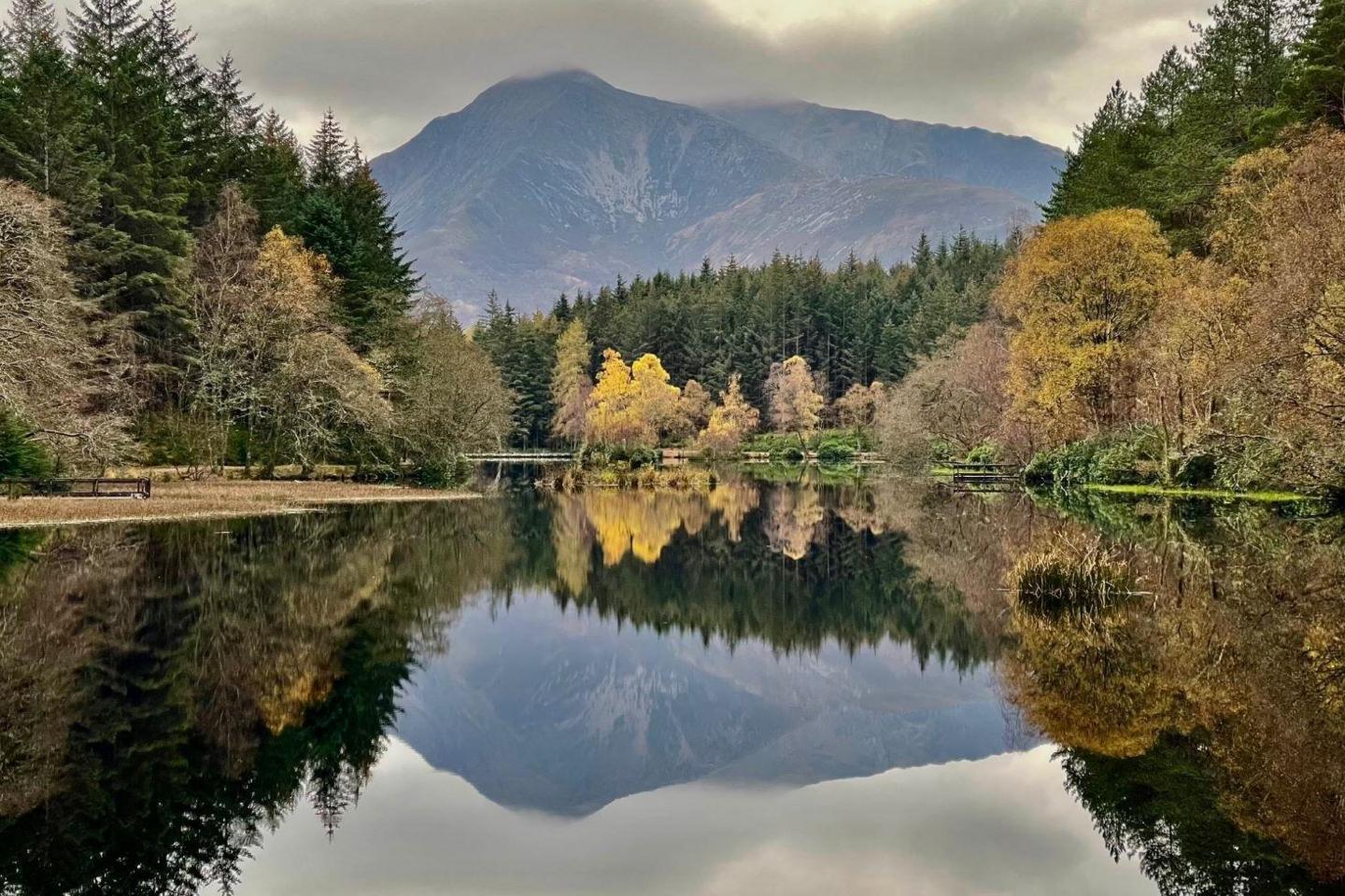 A mountain and trees are reflected in the loch. The trees have autumn colours include yellow and gold.